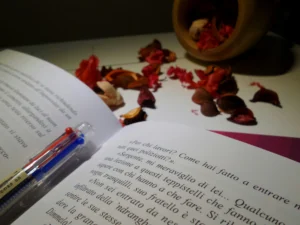 Open book with text in Italian, pen with red and blue ink lying on top. Wooden container spills vibrant potpourri onto a white surface, creating a serene, literary ambiance. Canadian Ghostwriters