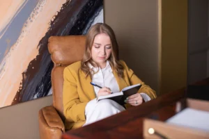 A woman in a mustard blazer sits in a leather chair, writing thoughtfully in a notebook. Abstract art adorns the wall, adding a creative ambiance. Canadian Ghostwriters