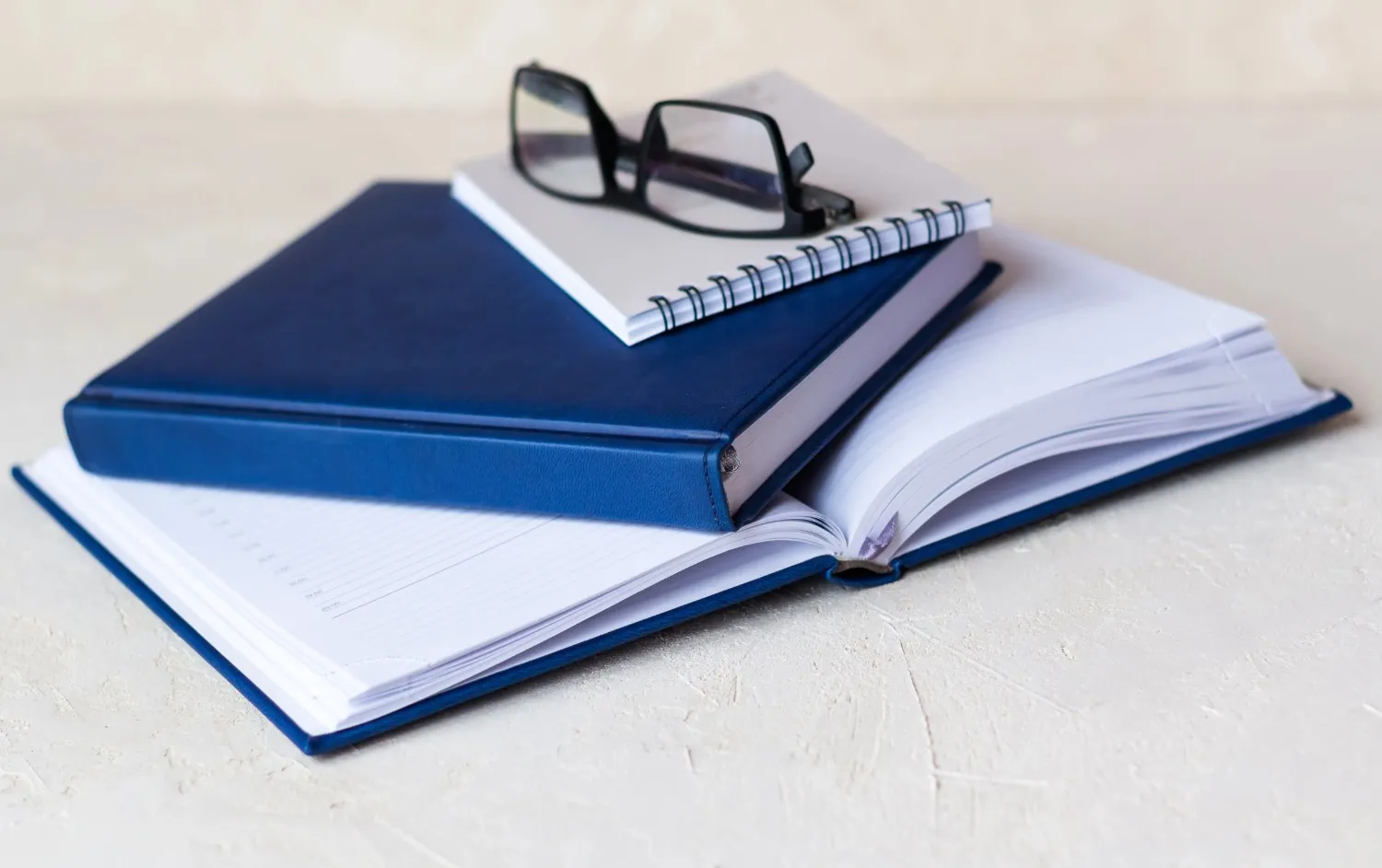 A pair of black-framed glasses rests on a stack of blue and white notebooks, with one blue notebook lying open on a light-colored surface. canadianghostwriters