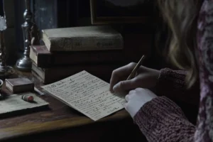 A person in a maroon sweater writes with a pen on a piece of cream-colored paper, sitting at a wooden desk with a stack of old, weathered books. canadianghostwriters