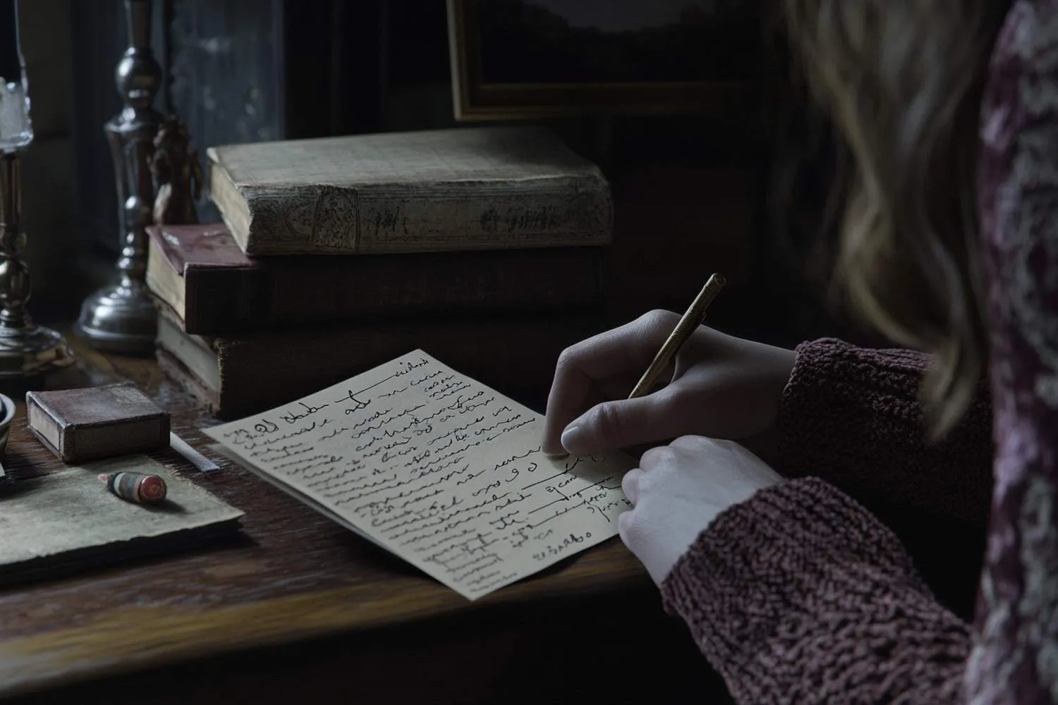 A person in a maroon sweater writes with a pen on a piece of cream-colored paper, sitting at a wooden desk with a stack of old, weathered books. canadianghostwriters