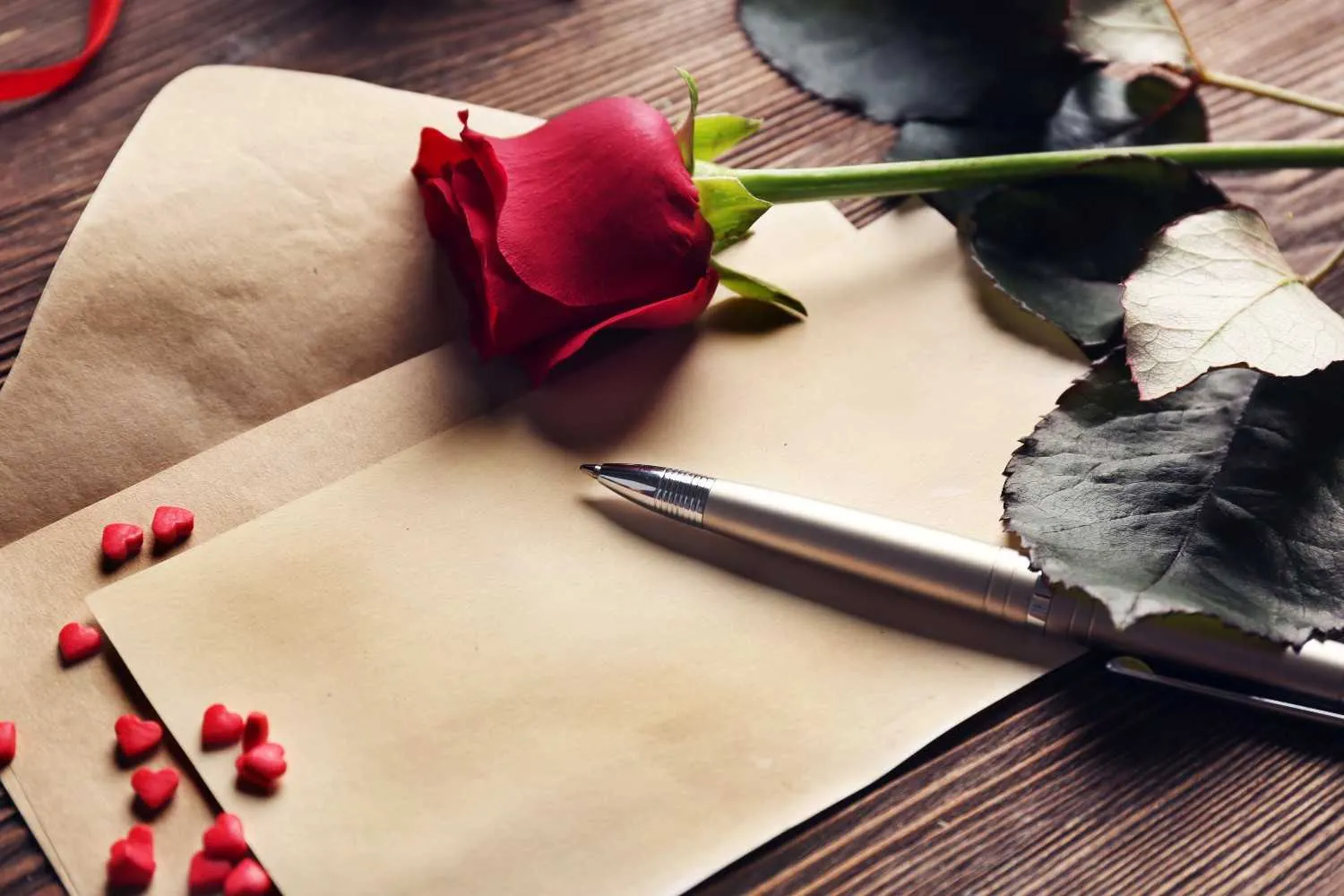 A single red rose, a metallic pen, and small red heart decorations resting on a blank beige envelope placed atop a dark wooden surface background. canadianghostwriters