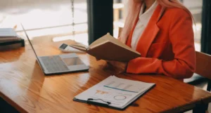 A person in an orange blazer reads a notebook at a wooden desk with a laptop, calculator, and clipboard holding papers with charts.canadianghostwriters