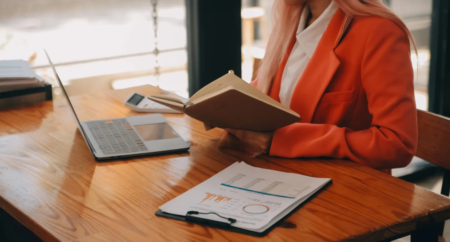 A person in an orange blazer reads a notebook at a wooden desk with a laptop, calculator, and clipboard holding papers with charts.canadianghostwriters
