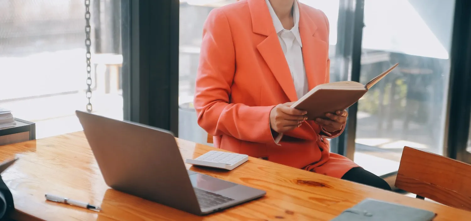 A person in a coral blazer reads a book at a wooden desk. A laptop and calculator are also visible on the desk. canadianghostwriters