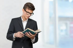A young man in a black suit and glasses is reading a book, standing indoors with large windows and a blurred background..canadianghostwriters