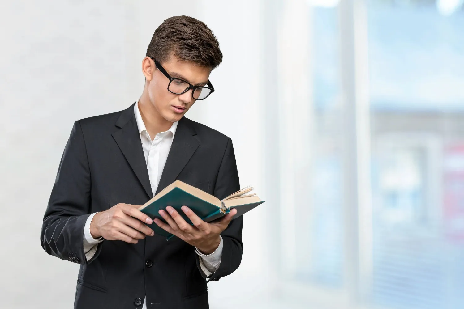 A young man in a black suit and glasses is reading a book, standing indoors with large windows and a blurred background..canadianghostwriters