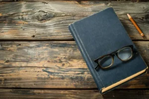 A closed blue book with black eyeglasses resting on top sits on a rustic wooden table next to a pencil. canadianghostwriters