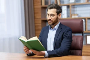A man in a suit reading a book while sitting at a desk.canadianghostwriters