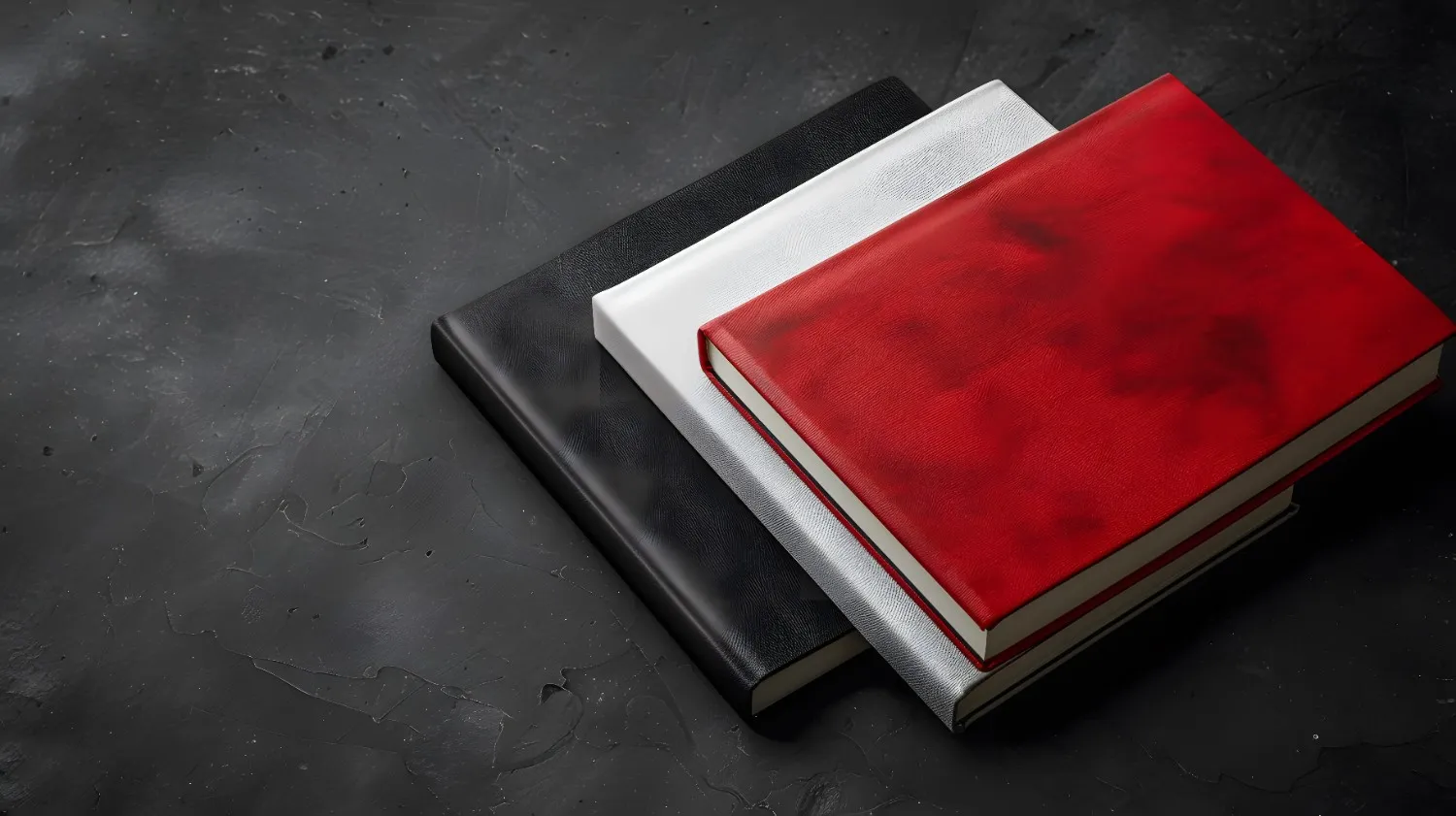 Three bound notebooks, black, silver, and red, are stacked on a dark textured surface. canadianghostwriters